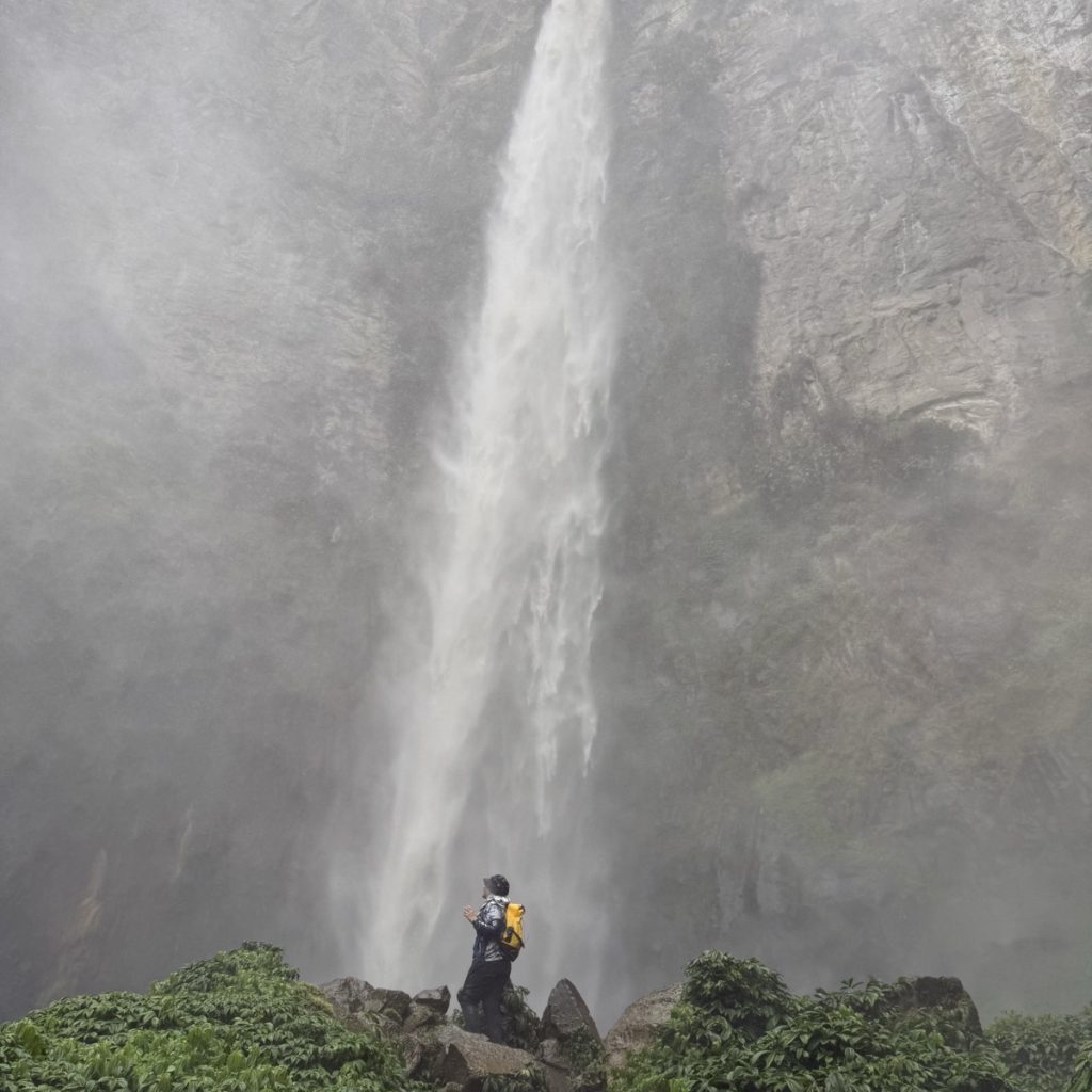 Tall waterfall in a misty forest with a hiker at its base
