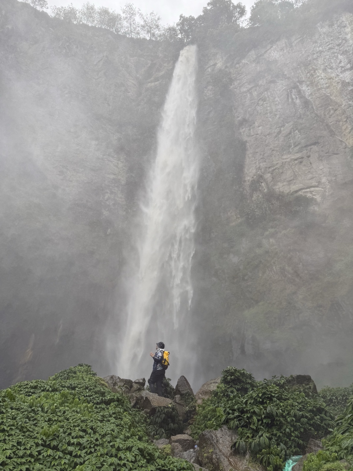 Tall waterfall in a misty forest with a hiker at its base